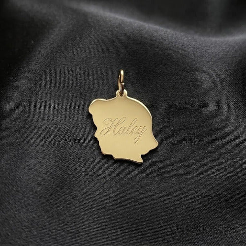 A gold-colored metal pendant charm shaped like a silhouette of a person's head, engraved with the name 'Haley' in cursive, resting on a black satin fabric background.