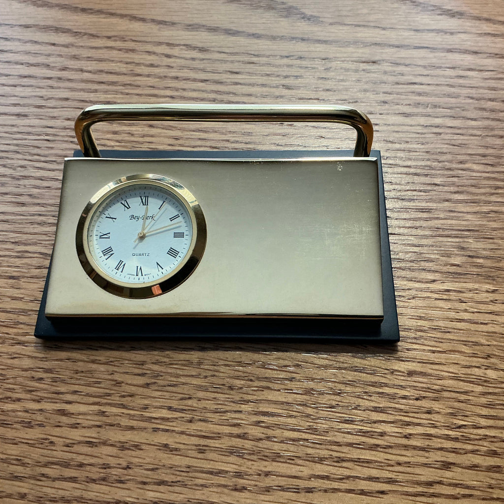 A vintage-style brass desk clock with a rectangular body, top handle, and round white dial, placed on a wooden tabletop.
