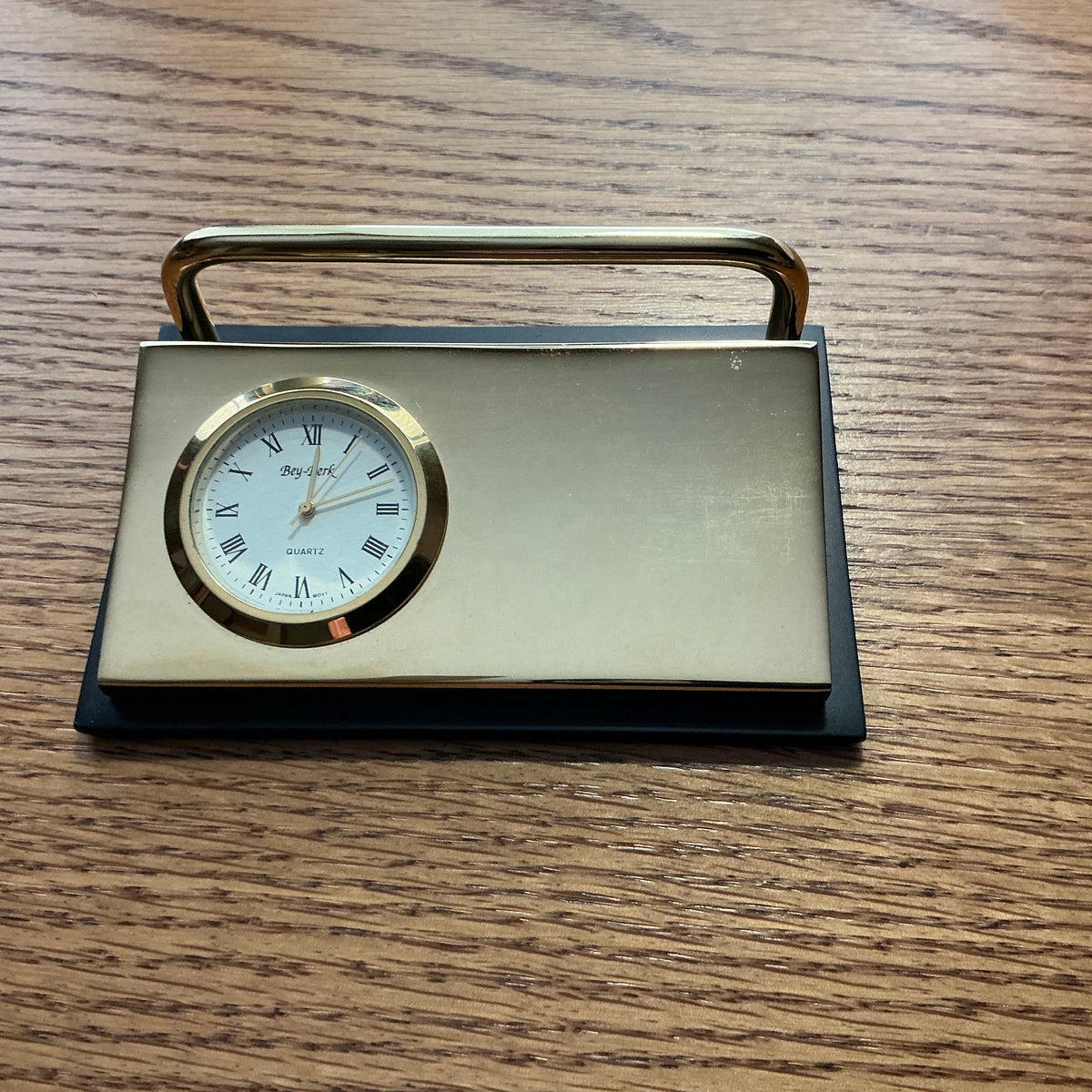 A vintage-style brass desk clock with a rectangular body, top handle, and round white dial, placed on a wooden tabletop.