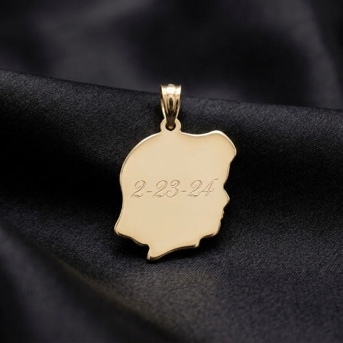 A gold pendant shaped like a profile silhouette with the date '2-23-24' engraved on its front, resting on a draped black fabric background.