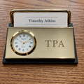 A personalized brass desk clock and name holder on a black base, featuring a round white clock face with gold trim, the engraved name 'TPA', and a printed card reading 'Timothy Atkins' on a wooden desk.