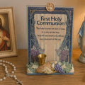 A First Holy Communion decorative plaque with printed religious text, blue floral borders, a small gold chalice, and a sculpted base with grapes, wheat, and leaves, displayed on a wooden surface with a rosary and framed religious image nearby.