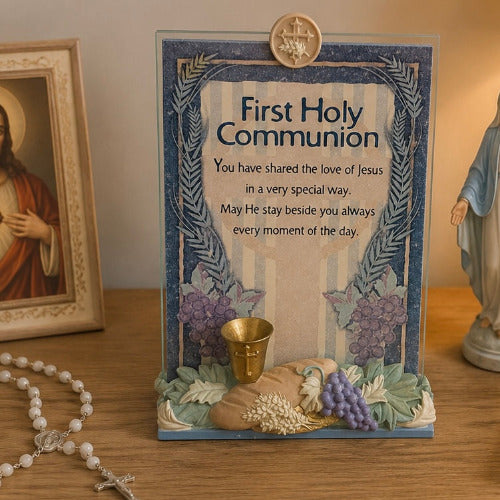 A First Holy Communion decorative plaque with printed religious text, blue floral borders, a small gold chalice, and a sculpted base with grapes, wheat, and leaves, displayed on a wooden surface with a rosary and framed religious image nearby.