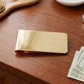 Gold money clip with initials on a wooden surface with money and a brush.