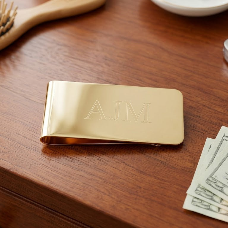 Gold money clip with initials on a wooden surface with money and a brush.