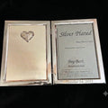A silver-plated brass photo frame shown open on a black background, with the left side displaying a heart-shaped engraving and the right side showing a sample photo insert with text including 'Silver Plated' and 'Bey-Berk International'.