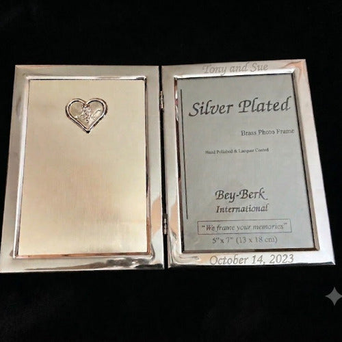 A silver-plated brass photo frame shown open on a black background, with the left side displaying a heart-shaped engraving and the right side showing a sample photo insert with text including 'Silver Plated' and 'Bey-Berk International'.