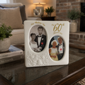 A white ceramic 60th wedding anniversary photo frame with two oval cutouts displaying wedding photos, set on a dark wooden coffee table in a cozy living room.