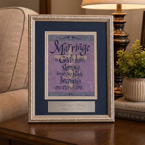 A framed scripture print reading 'Marriage is God's plan, strengthening one another' in purple and cream lettering, displayed on a wooden side table in a living room setting next to a lamp, a potted plant, and a sofa.