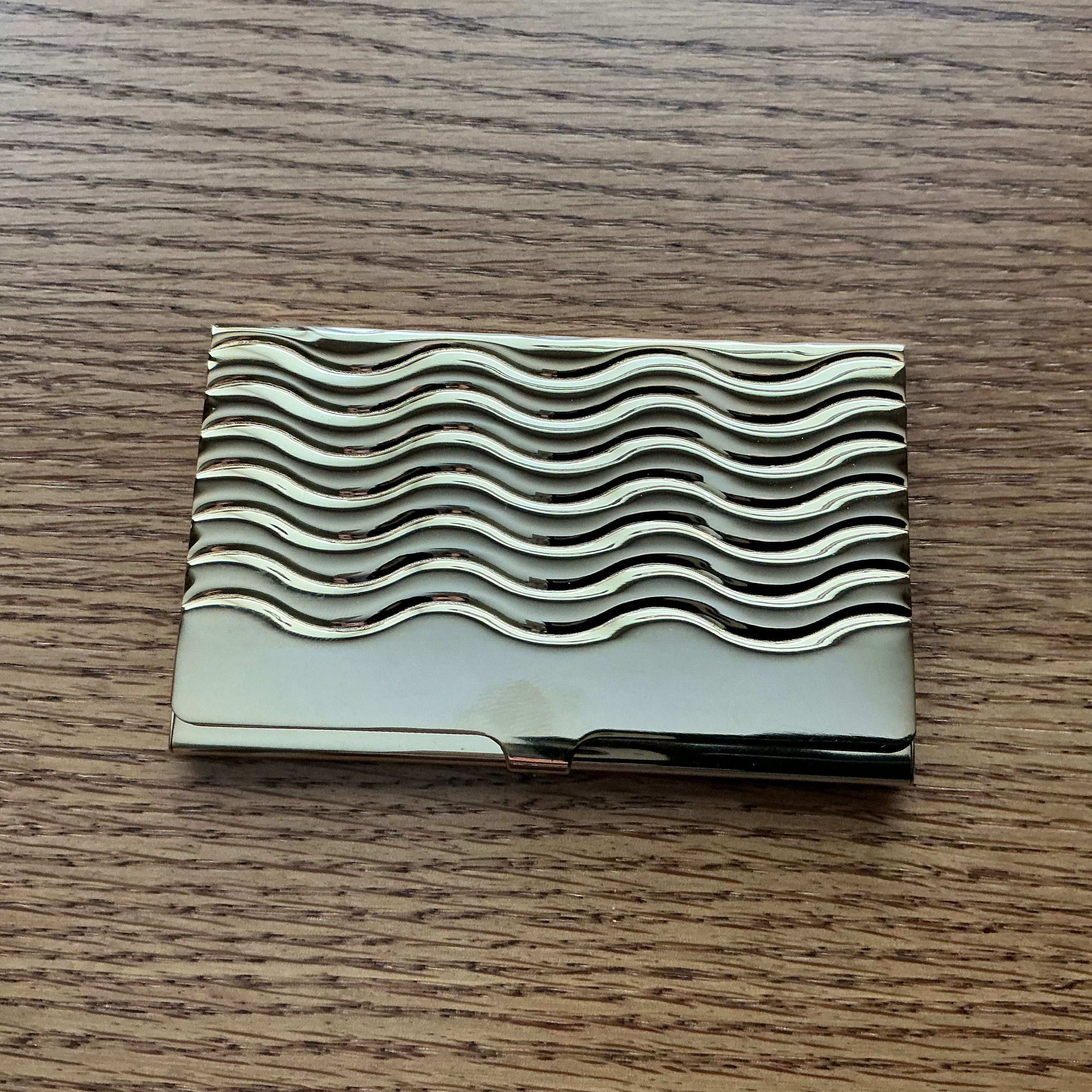A rectangular metallic business card case with a wavy, ridged pattern on the cover, resting on a brown wooden surface.