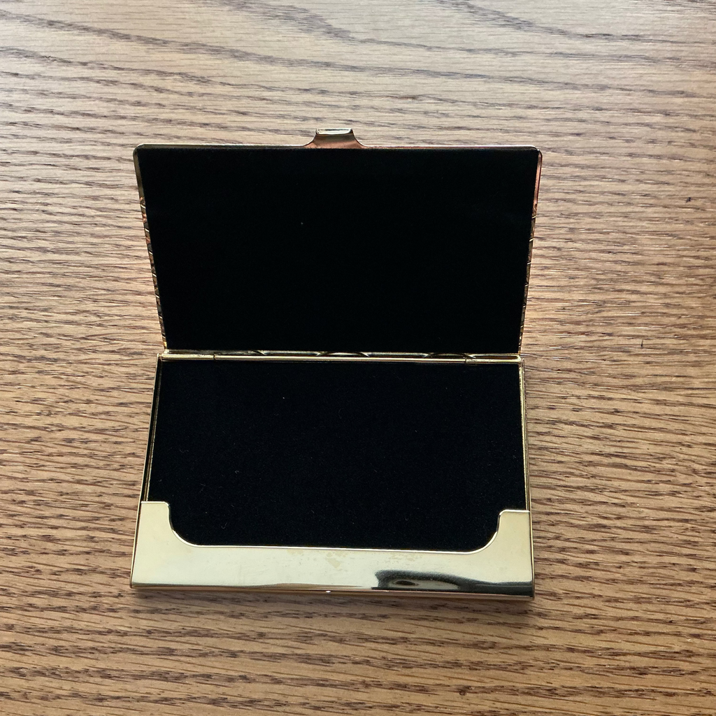 An open rectangular case with a black velvet interior and polished gold-tone metal trim, placed on a light brown wood-grain surface.