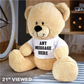 Beige teddy bear wearing a white shirt with 'ANY MESSAGE HERE' text, sitting on a wooden floor.
